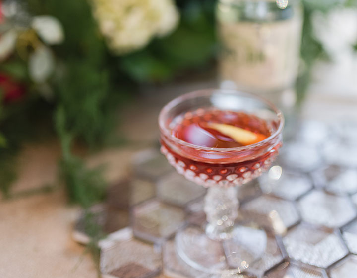 A blurry close up image of a pink cocktail in a fancy glass, a bottle of liquor in the background